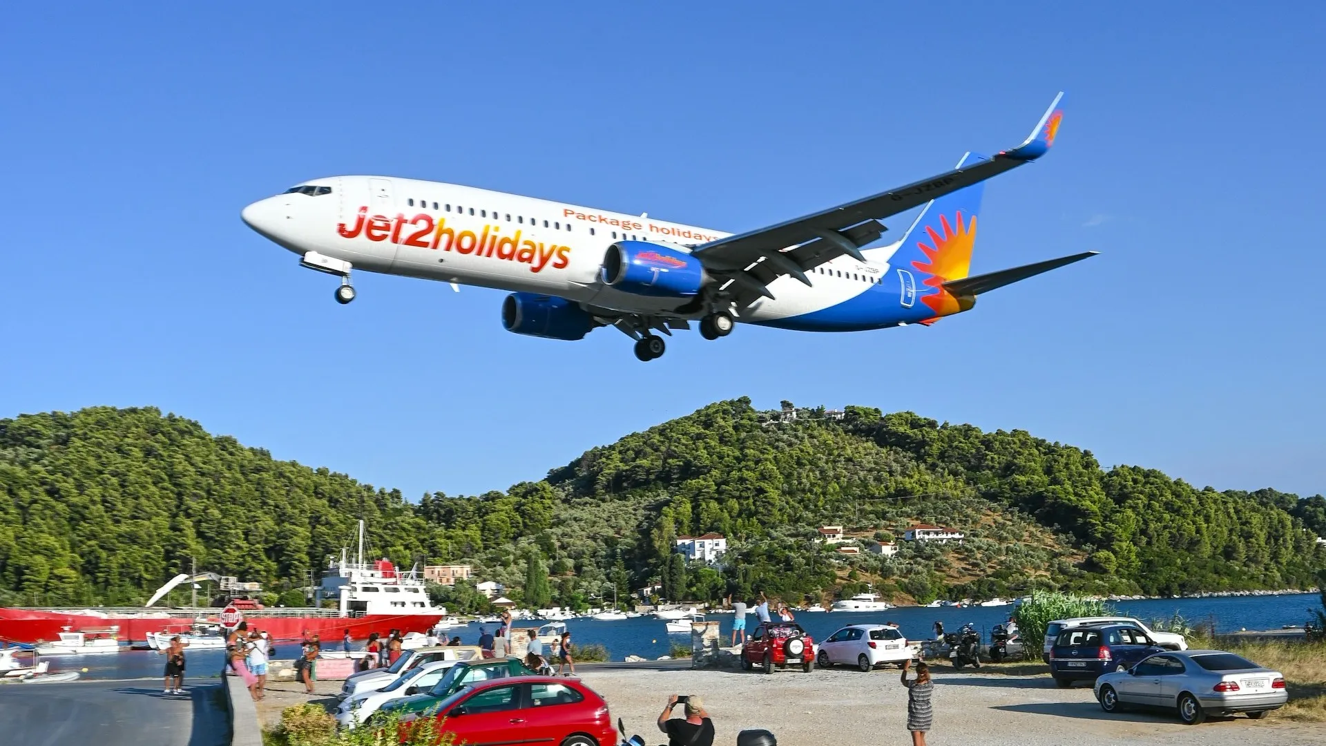 Samolot Jet2holidays podchodzi nisko do lądowania na Skiathos nad parkingiem z turystami i skuterami